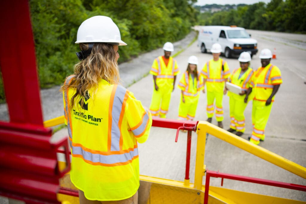 traffic plan crew members having a discussion while standing on a grey road next to line of green trees and bushes