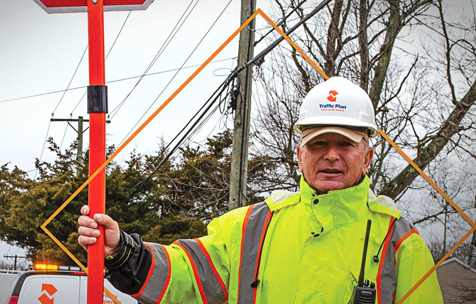 traffic plan employee holding stop sign while wearing cold weather company garb in front of trees and telephone pole