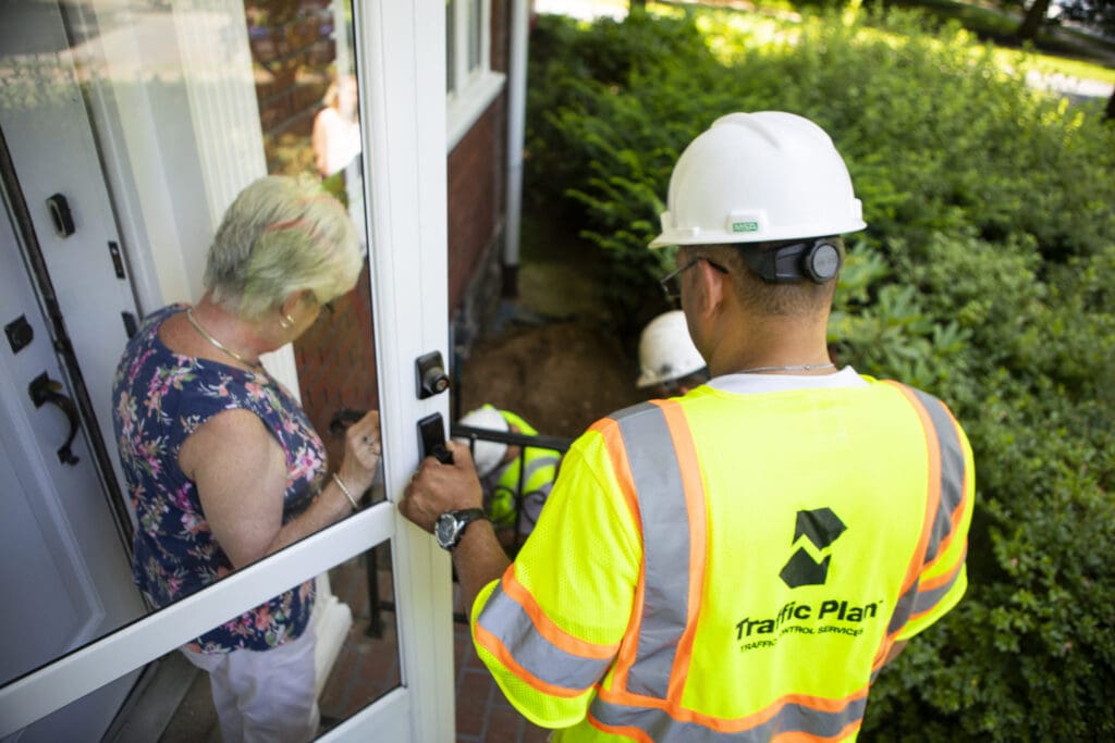 traffic plan flagger opening screen door for woman exiting home next to green shrubs
